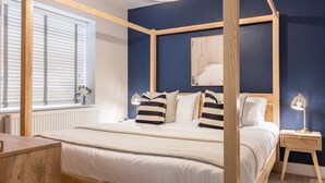 A bedroom with a four-poster bed, a bedside table with a lamp, a window with blinds, and a radiator. - Driftwood (Newquay)