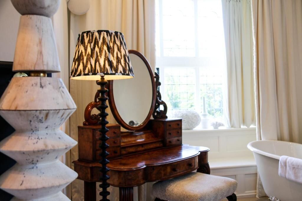 A vintage vanity with a mirror, a lamp, and a bathtub in a well-lit room with a window.