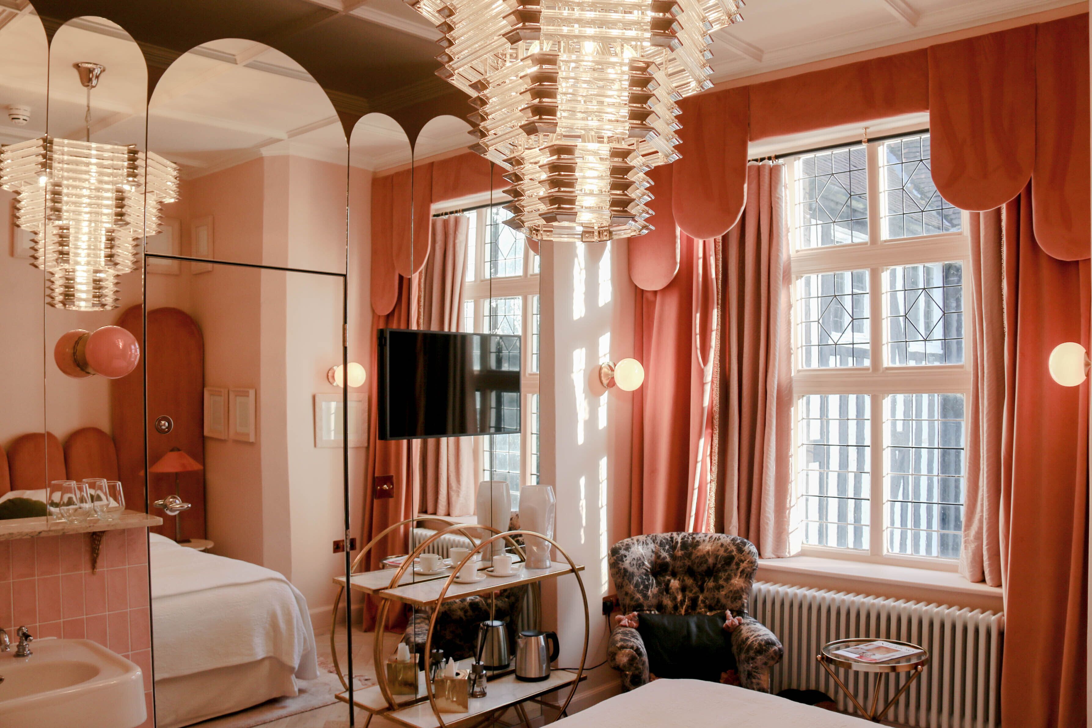 Habitación de hotel con un espejo grande, televisor, cama, tocador con lavabo y ventana con cortinas.