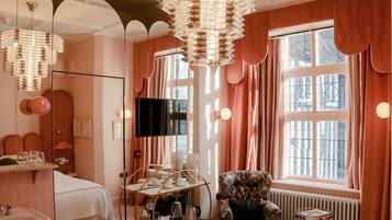 Habitación de hotel con un espejo grande, televisor, cama, tocador con lavabo y ventana con cortinas.