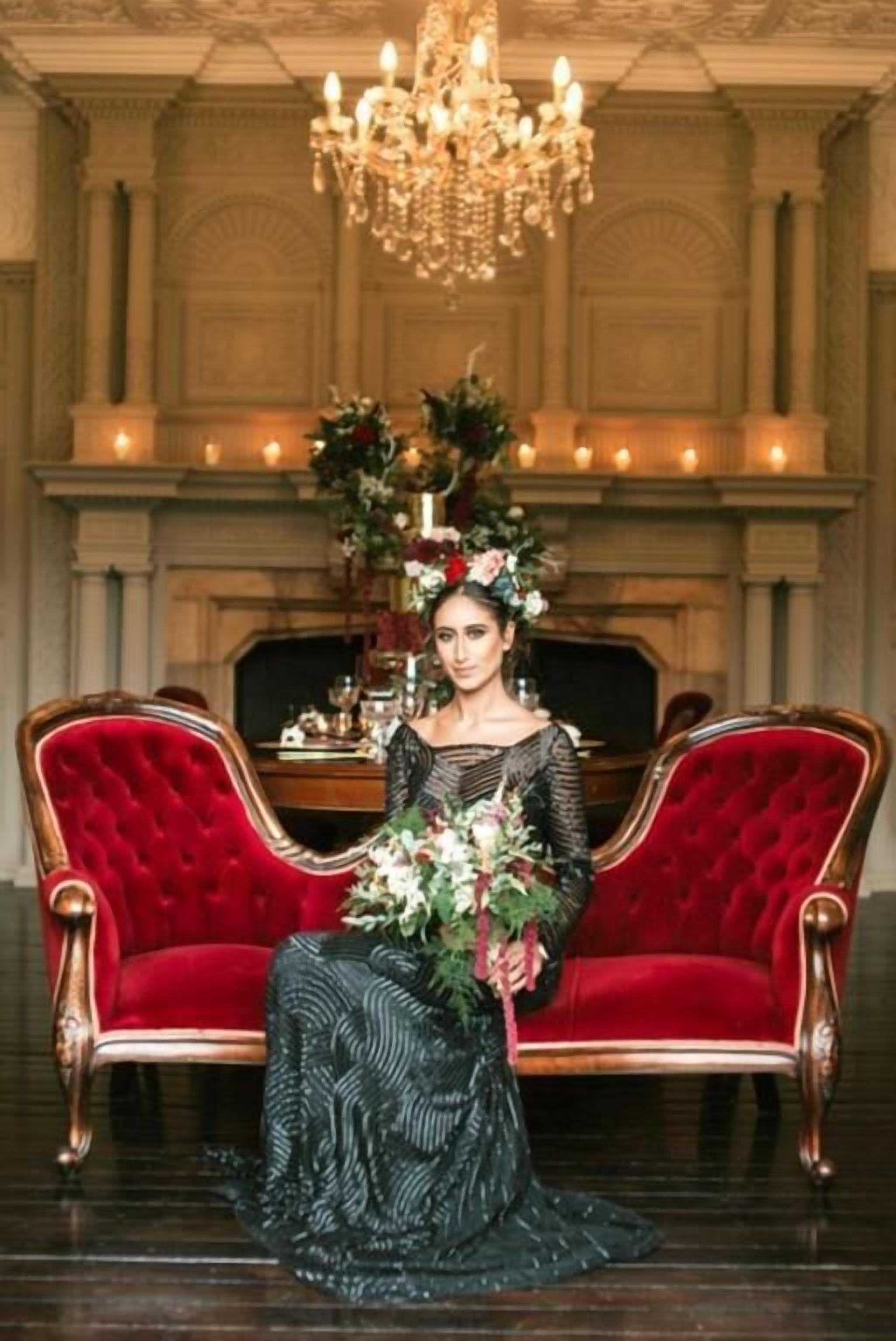 A woman seated on a red velvet sofa in a room with a fireplace, a chandelier, and a table with floral arrangements.