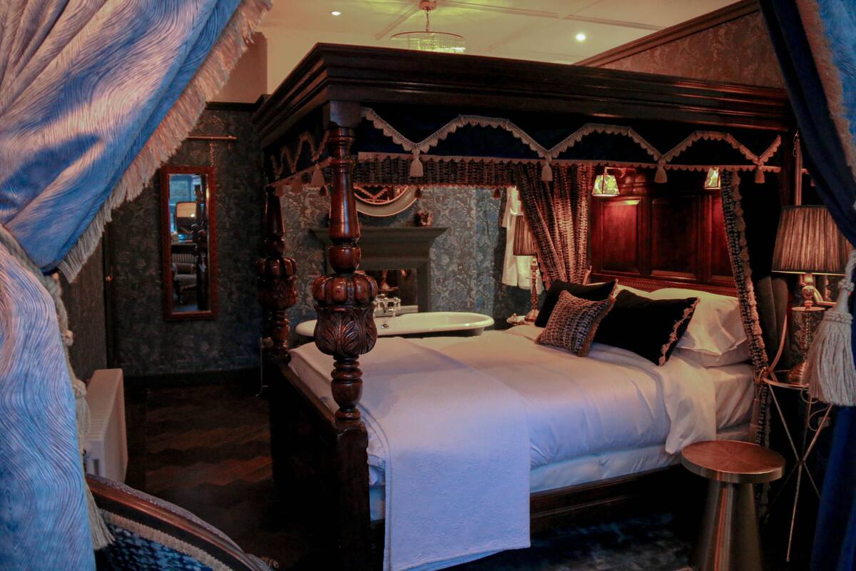 A four-poster bed with a canopy, a bedside table, a mirror, and a nightstand with a lamp.