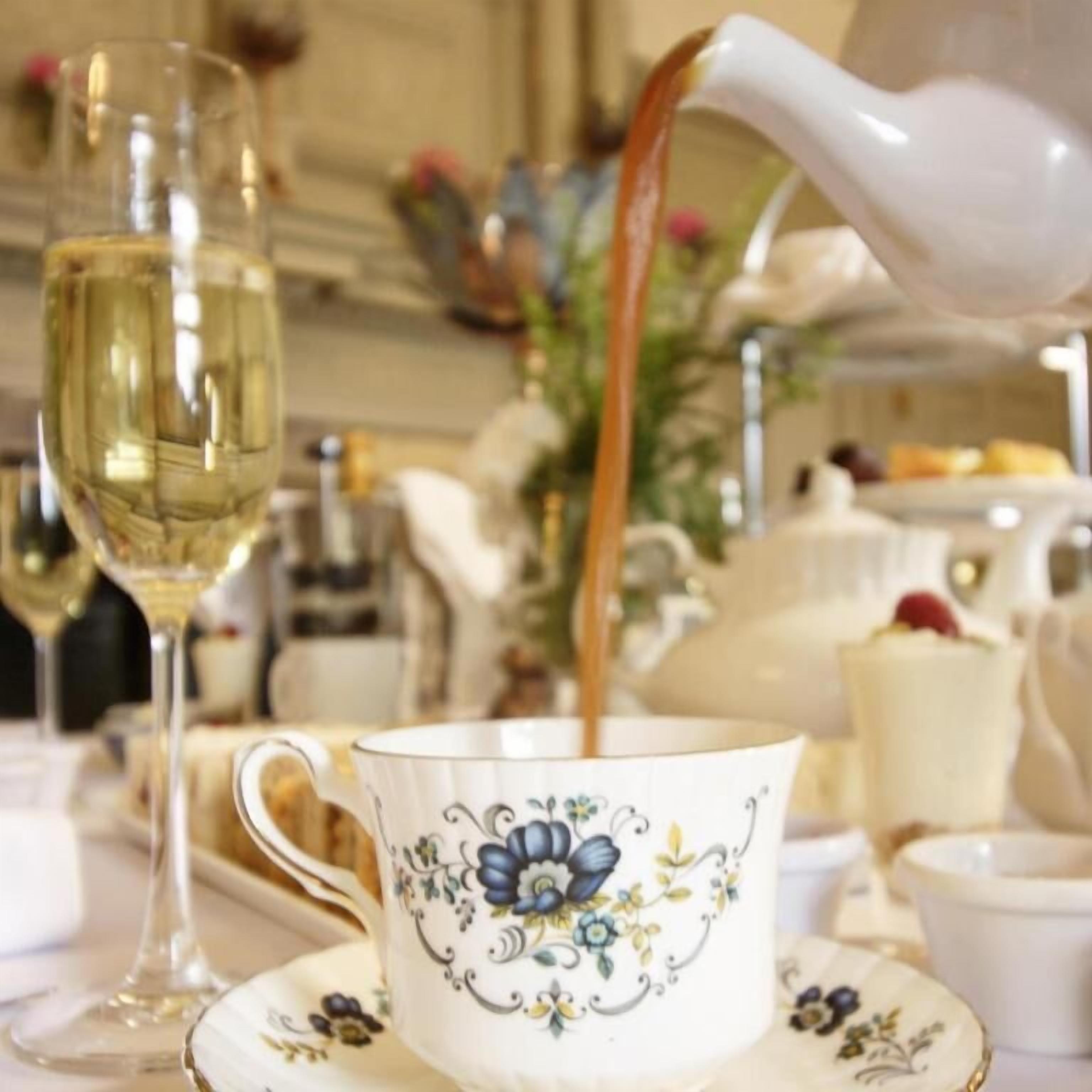 A table setting with a floral cup and saucer, a glass of white wine, and a teapot pouring tea.