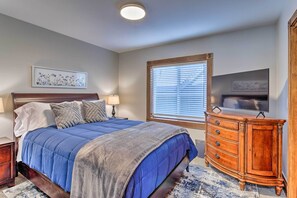 Une chambre à coucher comprenant un lit, une commode, une table de chevet, une lampe et une fenêtre avec des stores.