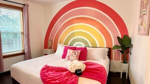 Ein Bett mit einem Kopfstück in Regenbogenfarben, einer rosa Decke und einem Kuscheltier.