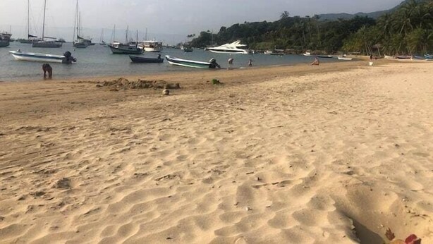 A sandy beach with boats docked in the background and a hilly landscape. - Casinha da Vila (Ilhabela)