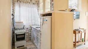 A compact kitchen with a stove, oven, and refrigerator, a small window, and a wooden shelf with items. - Casinha da Vila (Ilhabela)
