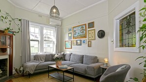 Una sala de estar con un sofá gris, una mesa de centro y una silla. Se observan cuadros en la pared, una ventana con cortinas y una lámpara colgante.