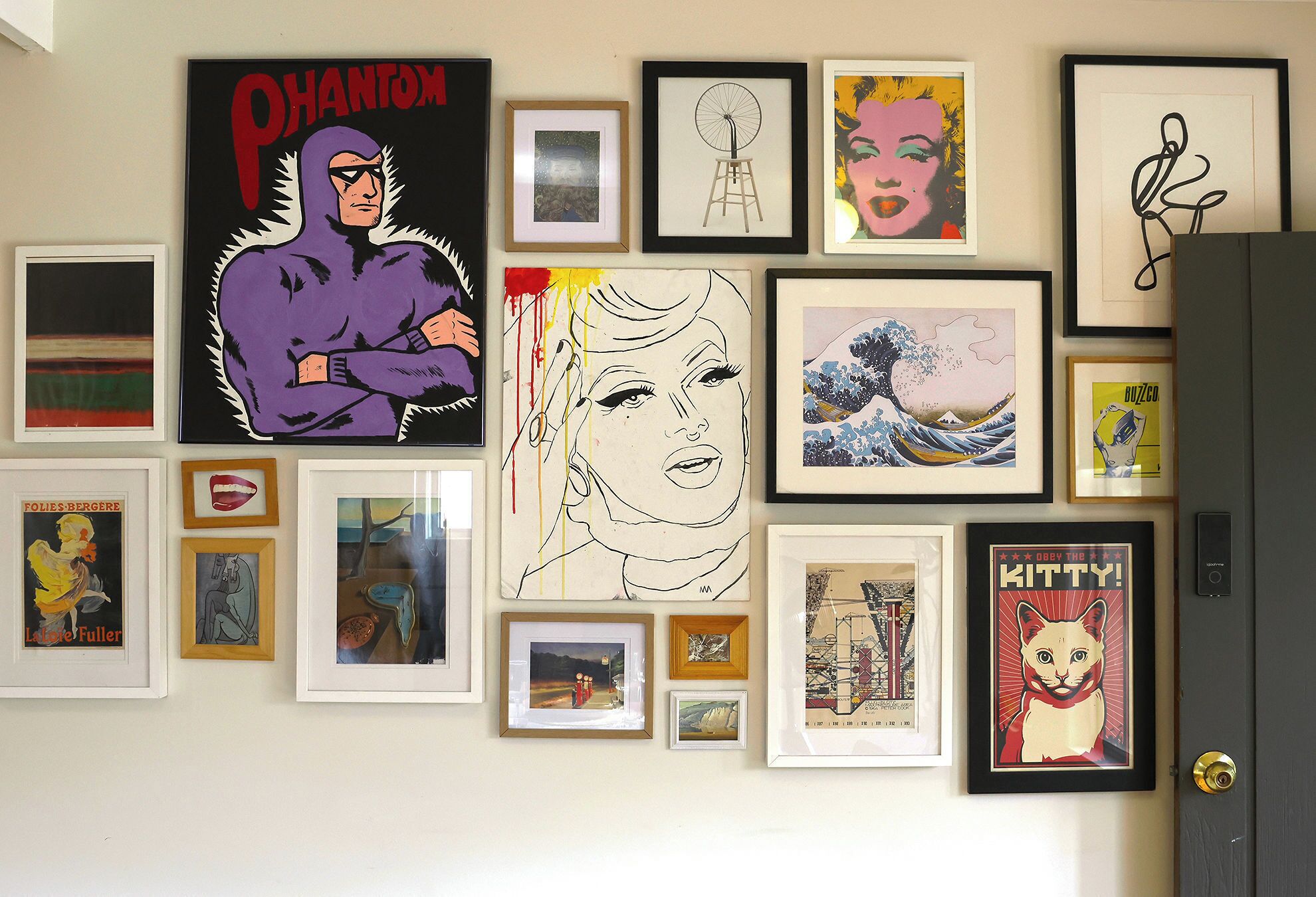 Una parete con diverse opere d'arte incorniciate, tra cui un poster di Phantom, un ritratto di donna e un'illustrazione di un gatto.