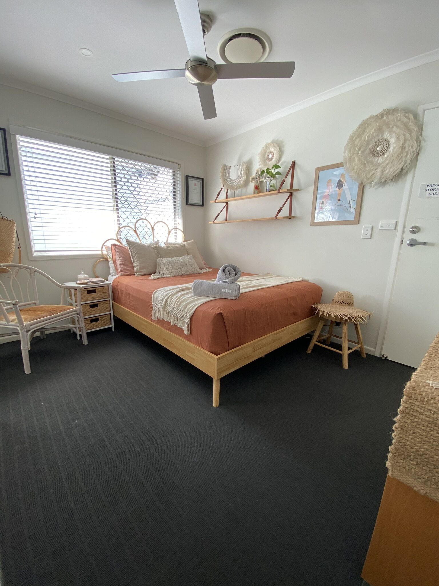 Ein Schlafzimmer mit Bett, Deckenventilator, Fenster mit Jalousien, Nachttisch, Stuhl und einem kleinen Hocker.