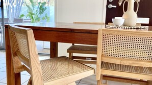 Un tavolo da pranzo in legno con una sedia in fibra intrecciata e una sedia in rattan, un'opera d'arte incorniciata al muro e un vaso con fiori secchi su un tavolo.
