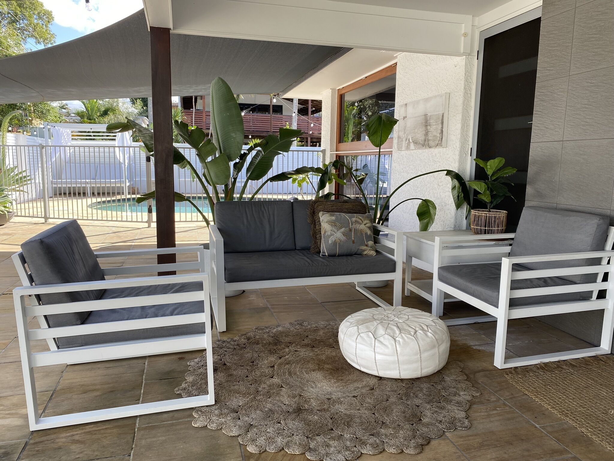 A covered outdoor seating area with white furniture, a woven rug, and large plants.
