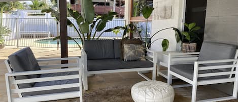 A covered outdoor seating area with white furniture, a woven rug, and large plants.
