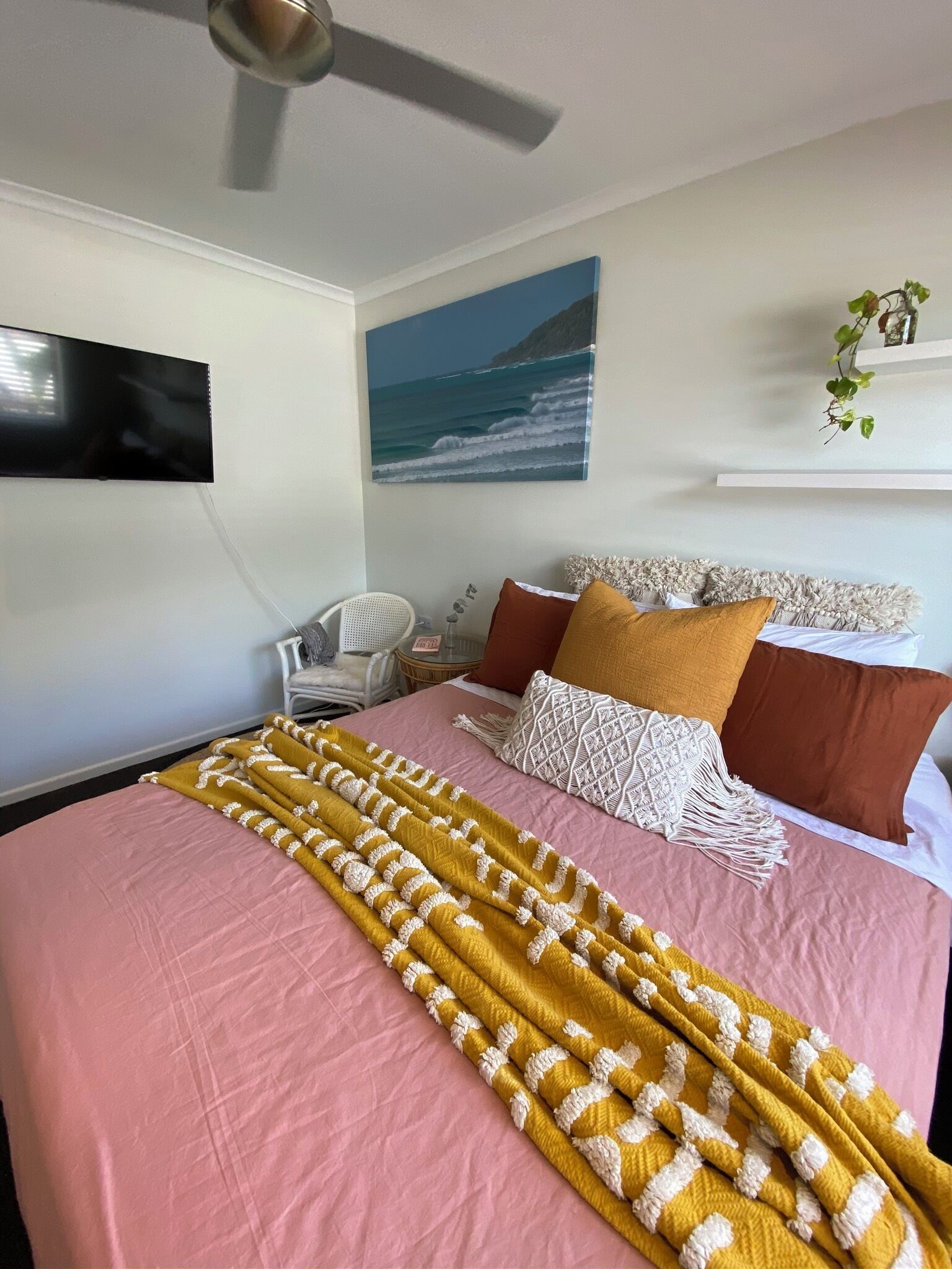 Une chambre à coucher avec un lit, des oreillers, une couverture, un ventilateur de plafond, un téléviseur fixé au mur et un tableau représentant l’océan.