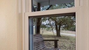 Eine Holzbank mit einer Metall-Schaukel, vor einem Fenster mit Blick auf eine Wiese und Bäume.