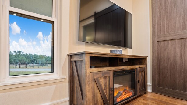 Ein Wohnzimmer mit großem Fernseher über einem Holzschrank mit Glastür, einem Kamin und Blick auf einen von Bäumen gesäumten Garten durch ein Fenster.