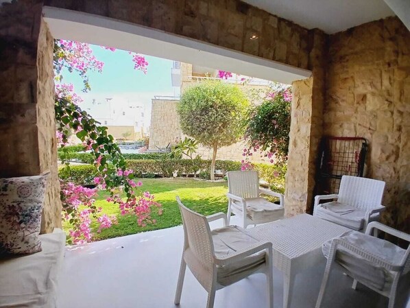A patio with white wicker furniture, a stone wall, and a view of a garden with pink flowers and a tree. - El Gouna Swan Lake on the shores of the Crystal Lagoon, conference room, 10 minutes from downtown, marina (Hurghada Al-Bahr al-ahmar)