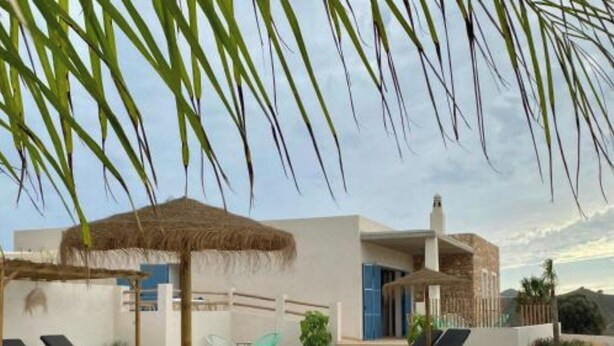 Un'area piscina moderna con sedie a sdraio, un ombrellone di paglia e un edificio con rivestimento in pietra.