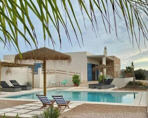 Un'area piscina moderna con sedie a sdraio, un ombrellone di paglia e un edificio con rivestimento in pietra.