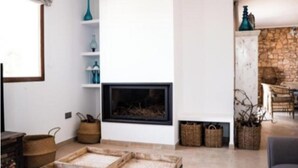 Un soggiorno con camino, pareti bianche e una parete in pietra sullo sfondo. Sono presenti mensole con oggetti decorativi e una finestra che lascia entrare luce naturale.