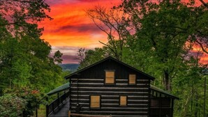 A rustic log cabin with a porch, surrounded by lush greenery and set against a vibrant sunset sky. - Pet Friendly · Hot Tub · Mins to Dollywood · Views (Pigeon Forge)