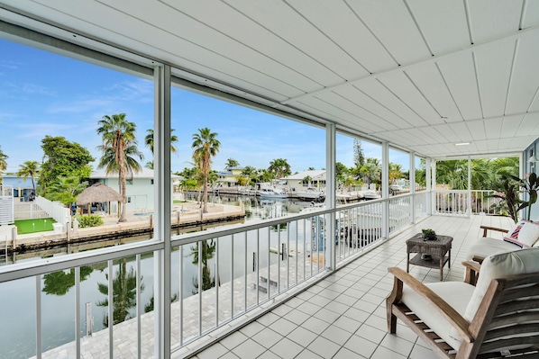 A covered balcony with a view of a marina, palm trees, and a pool.