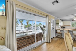 A room with a large sliding glass door leading to a balcony, a wooden bench, a kitchen area with a toaster, and a view of a marina with boats and palm trees.