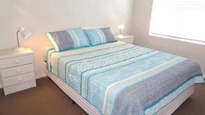 Un letto con una coperta a motivi, due lampade ai comodini, un comodino bianco e una finestra con persiane.