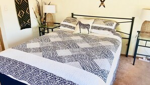 A bed with a zebra-patterned quilt, a bedside table with a lamp, and a chair with a woven basket. - The Peninsula - Lifestyle Yamba (YAMBA)