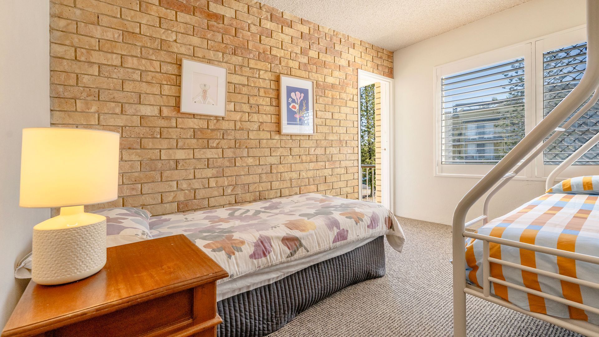 Un dormitorio con pared de ladrillo, una cama con colcha floral, una mesita de noche con lámpara y una ventana con persianas.