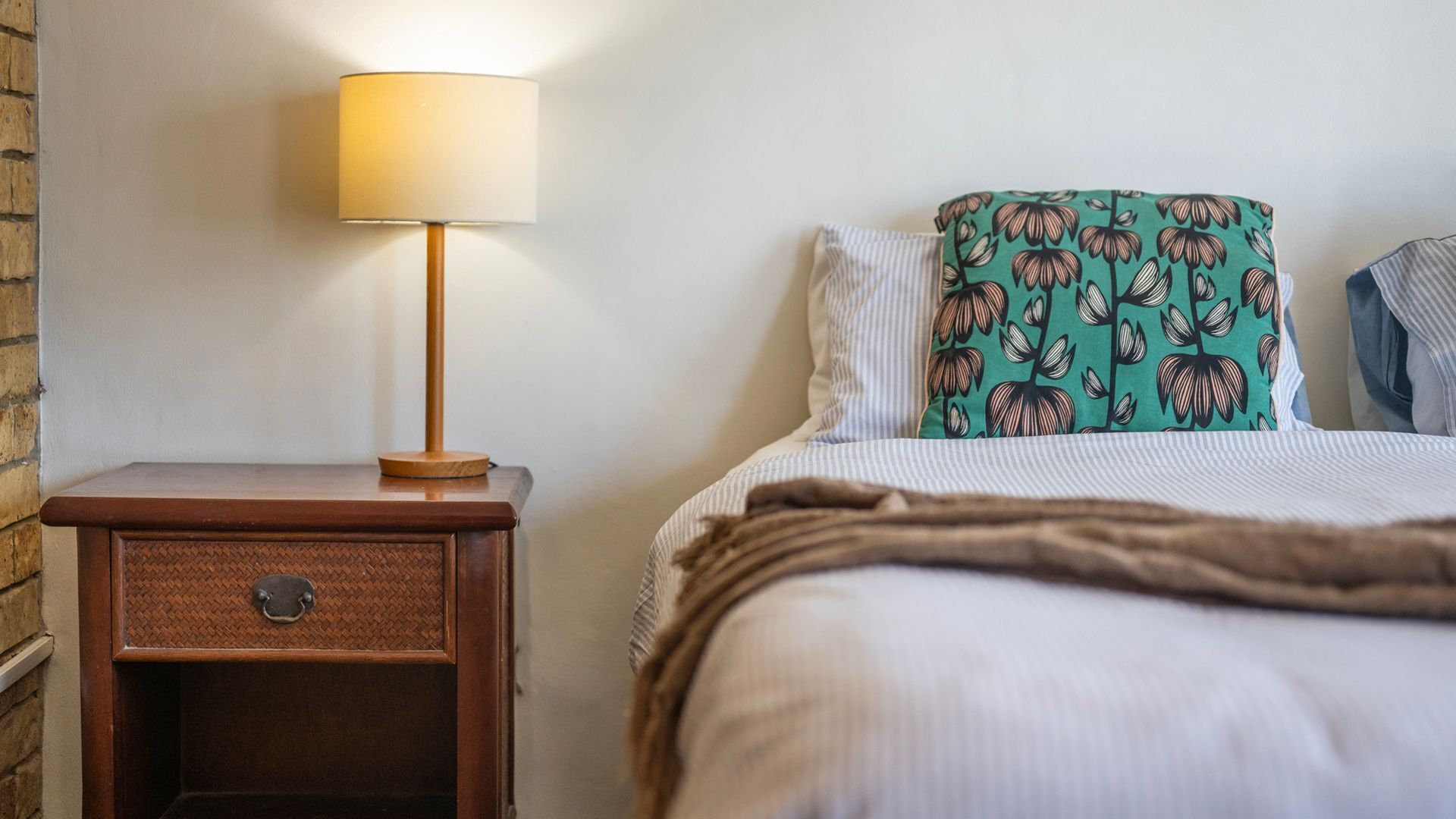Een net opgemaakt bed met een gestreept kussen en een bloemenkussen in groen, een bedlamp op een nachtkastje van hout, en een baksteenmuur.