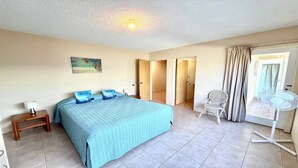 Une chambre à coucher comprenant un lit, une table de chevet, une chaise, un ventilateur et une porte donnant sur une autre pièce.