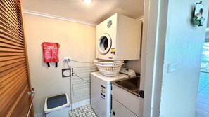 A compact laundry area with a washing machine, a drying rack, a fire blanket, and a small trash bin.