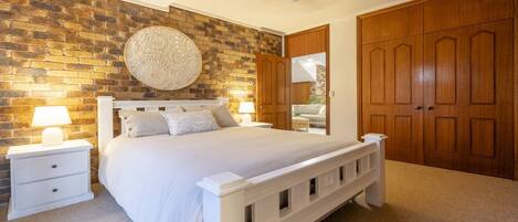 A bedroom with a brick wall, a round decorative wall piece, a wooden door, a white bed with pillows, and two bedside tables with lamps.