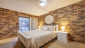 A bedroom with a brick wall, a round decorative wall piece, a bed with white bedding, two bedside tables with lamps, and a window with a view.