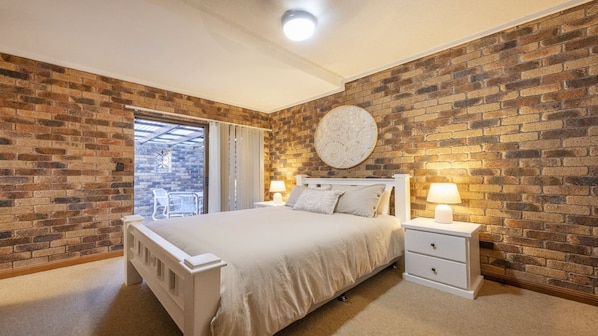 A bedroom with a brick wall, a round decorative wall piece, a bed with white bedding, two bedside tables with lamps, and a window with a view.