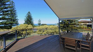A wooden deck with a table and chairs, overlooking a coastal landscape with trees and a clear sky.
