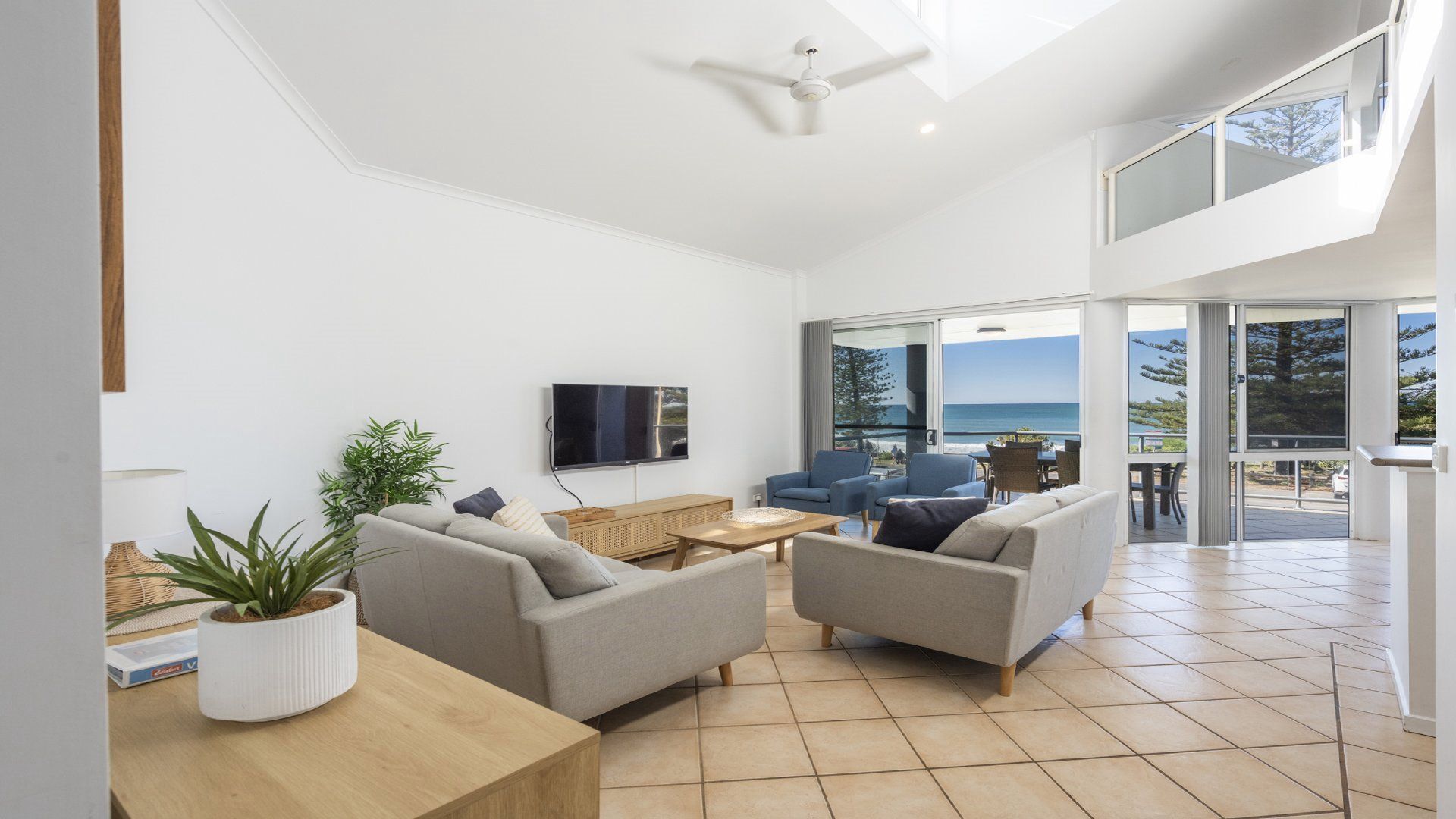 A modern living room with a flat-screen TV, two grey sofas, a wooden coffee table, and a large window offering a view of the sea.