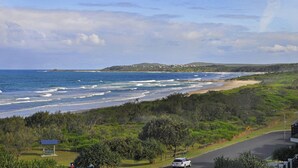 A coastal road with a bench and a car, leading to a beach with waves and a hilly landscape in the background. - Breakers Block 2 Unit 11 - Lifestyle Yamba (YAMBA)