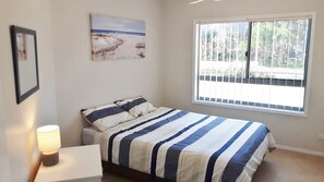 Una camera da letto con un letto, un comodino, una lampada, una finestra vista alberi e un quadro appeso al muro.