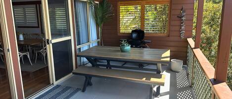 Un patio al aire libre con mesa y sillas de madera, una parrilla y una ventana con persianas.