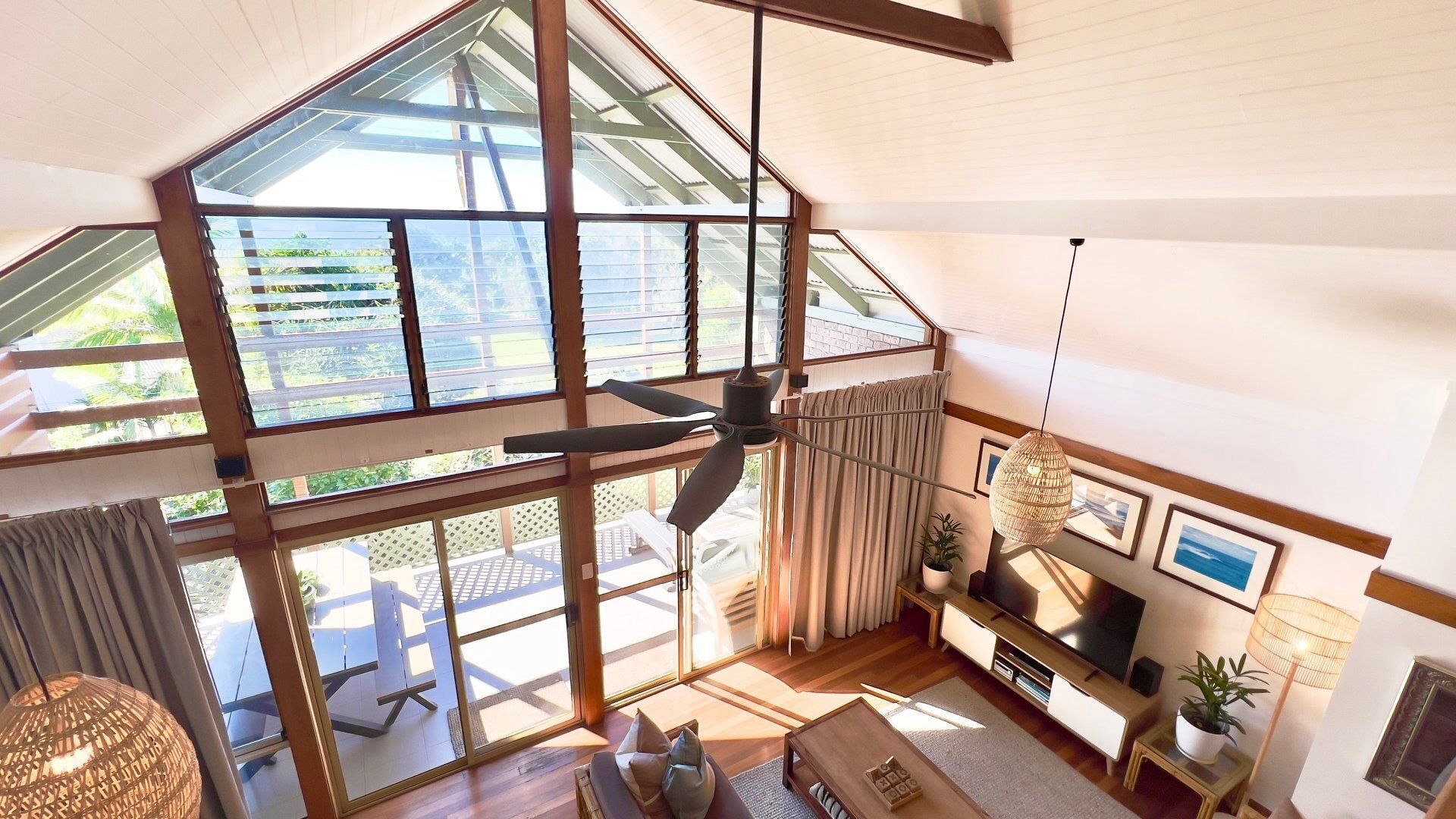 A spacious living area with a high ceiling, large windows, a ceiling fan, and a television.