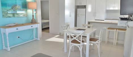 Een moderne keuken met witte kastjes, een eethoek met witte stoelen en een televisie op een standaard.