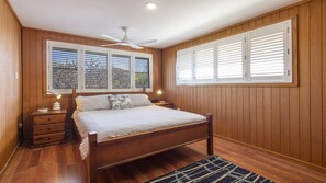 A bedroom with a wooden bed, two bedside tables, a ceiling fan, and large windows with shutters.