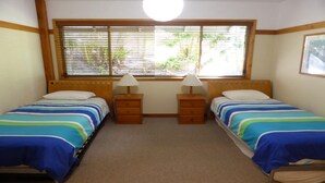 A room with two single beds, each with a wooden headboard and a bedside table with a lamp. There is a large window with blinds, and a ceiling light fixture. - Drift - Lifestyle Yamba (ANGOURIE)