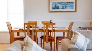 A dining area with a wooden table and chairs, a framed picture on the wall, and a window providing natural light. - Meridian 5 - Lifestyle Yamba (YAMBA)