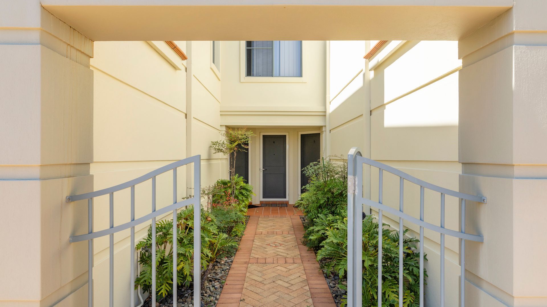 A narrow pathway leading to a building entrance with a door and window, surrounded by greenery and a brick pathway.