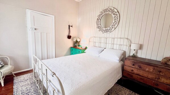 A bedroom with a white bed, a wooden dresser, a green nightstand, a white chair, and a wall-mounted mirror.