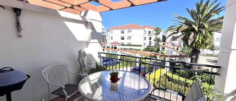A balcony with a glass table, two chairs, a statue, and a view of buildings and palm trees.