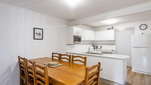 A compact kitchen with white cabinetry, a wooden dining table with chairs, a microwave, a sink, and a refrigerator. - Ocean Spirit - Lifestyle Yamba (YAMBA)